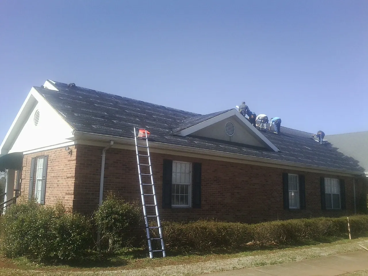 Crew tearing shingles off a brick ranch roof with a ladder set — Metal Roof Repair work by our crew in Alleyton