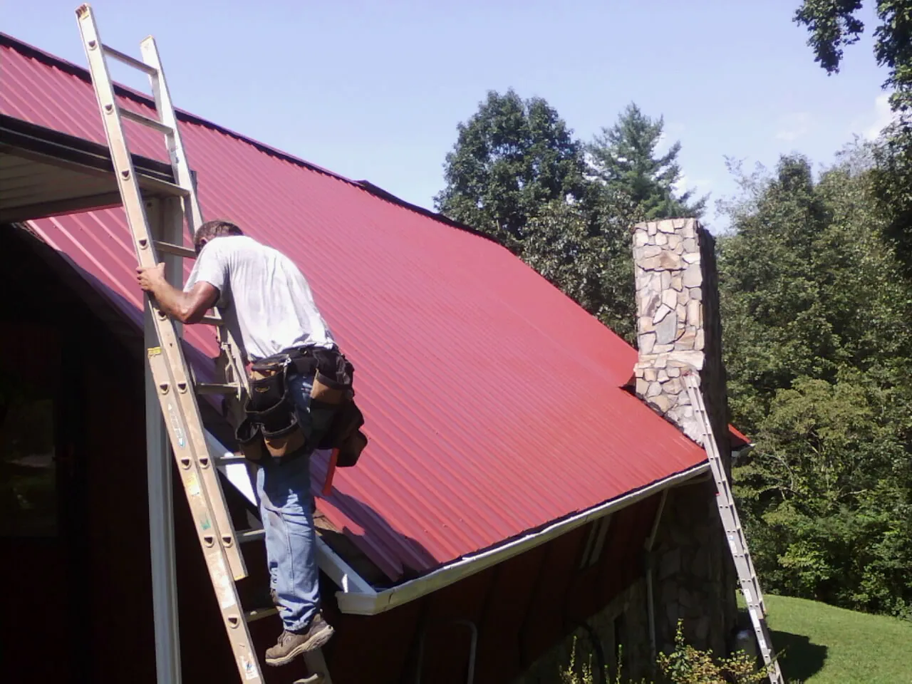 Roofer on a ladder beside a red metal roof and stone chimney — Soffit & Fascia Repair by Roofing Brothers in Alleyton