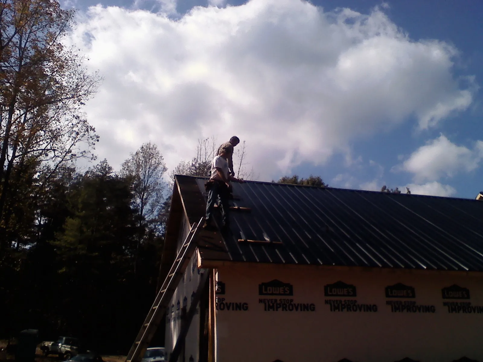 Roofer fastening dark metal panels high against a cloudy sky, from a Roof Inspection job in Alleyton